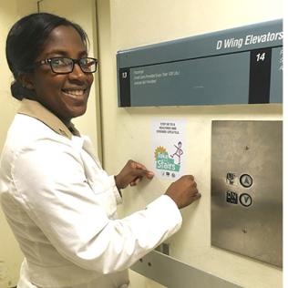 Holshue, in a white lab coat, stands in front of Take the Stairs sign next to elevator in the Clinical Center