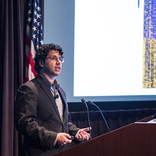 Goodarzi stands with cupped hands at podium in front of purple, yellow presentation slide.