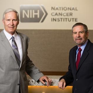 Schiller, Lowy stand in front of National Cancer Institute sign.