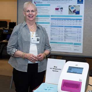 Gaydos stands beside diagnostic instrument, with scientific poster behind her.