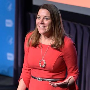 Karin Hurt in red dress stands on stage with hand extended.