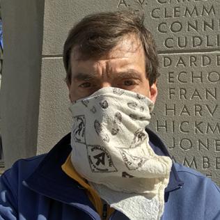 Professor Ewing, wearing a cloth mask, stands in front of pylon etched with names of World War I victims