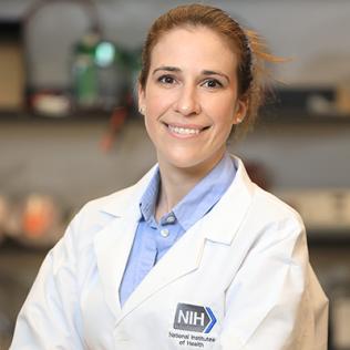 A smiling Sadtler, in white lab coat with NIH logo, standing in the lab