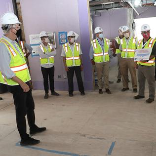 About a dozen people in helmets, masks, and safety vests stand in an unfinished room as one speaks.