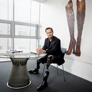 Herr is smiling, seated at a table. His prosthetic legs are prominent. On a wall behind him is an artwork of a pair of legs.
