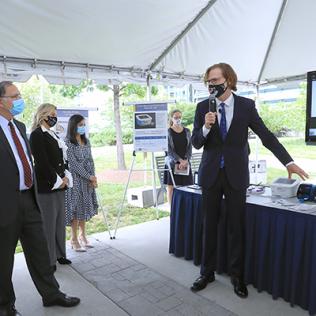 Tromberg stands in front of a display table and speaks to several senators under a tent