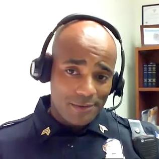 screenshot Black male police officer wearing uniform and headset, speaks into computer screen