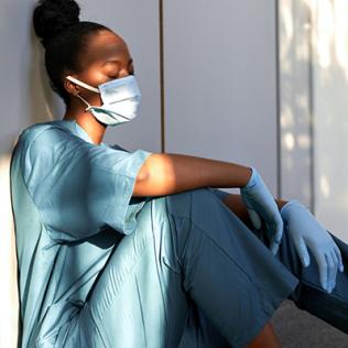 A female nurse in blue scrubs, mask and gloves sits, leaning against a hospital wall, asleep.