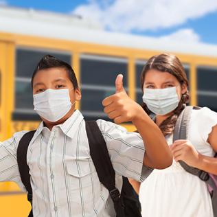 Three Latino kids, wearing backpacks and smiling behind their masks, give thumbs up - in front of a school bus with a bright blue sky overhead.
