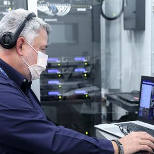 Burnham sits in office with 2 laptops on desk and computer servers and other equipment behind him.