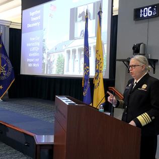 Levine speaks at an NIH podium, with event title slide projected on screen beside her