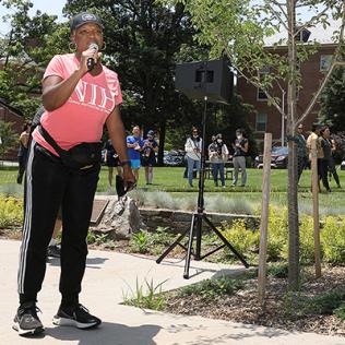 Edwards in pink NIH shirt talks into microphone as dozens of hikers stand in front of building 1 waiting for hike to begin.