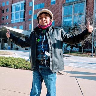 Caesar stands smiling with arms extended and both thumbs up in front of the Clinical Center during a recent checkup.