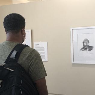 A man wearing a backpack reads a narrative on the wall next to portrait drawn by Clower. The exhibit is displayed on the main floor, east alcove, of the Clinical Center.