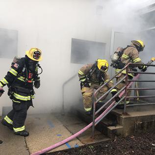 Three fire fighters in uniform walking into smoky building with hose on NIH campus, as part of a training exercise