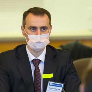 Ukraine's health minister wearing a mask, sits with hands folded at table in CC medical board room, listening intently to NIH staff briefing.