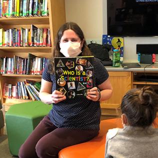 Gehl sits in the Inn lounge, surrounded by books, holding up her book as kids look on.