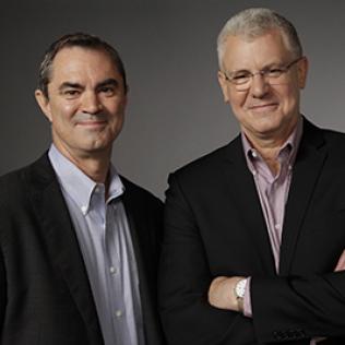 Smith and Johnson smile shoulder-to-shoulder. Smith, left, wears a black suit jacket and blue shirt, and Johnson wears glasses, a black suit jacket and checkered red and white shirt.