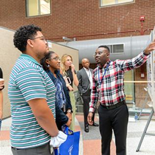 Man in plaid shirt explains his research poster to onlookers