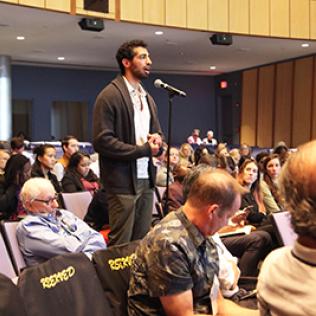 Man in brown cardigan speaks at microphone in auditorium