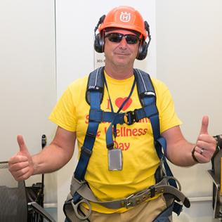 Man in yellow Wellness day shirt, cap, sunglasses, headphones and safety harness gives thumbs up.