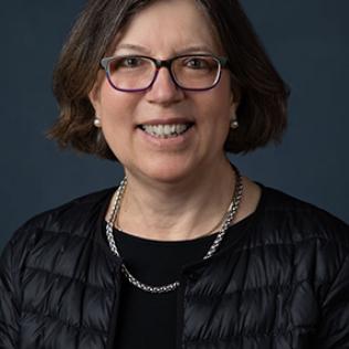 Sears smiles against a dark blue-gray background. She wears glasses and a black jacket.