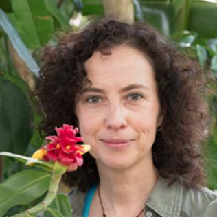 Vandebroek smiles softly in front of a leafy green plant. A red flower extends close to the right side of her face.