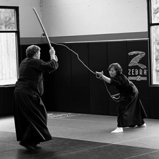 B&W photo of man in martial arts pants and tunic holds up rod facing woman in similar uniform, slightly crouching, holding up rod and rope. 