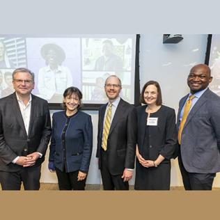 Three men and two women stand smiling in front of projected slide of All of Us research sample faces from multicultural dataset