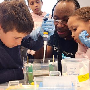 Kid holds pipette as Dr. Manu Platt guides his arm, and other kids look on.