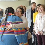 A Native American woman in a colorful sweater hugs a colleague as others smile, looking on.