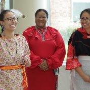 Three women in traditional Native garb stand together, smiling.