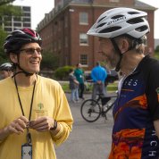 Two smiling cyclists wearing helmets stand outside discussing biking to work.