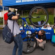 In front of NIH Police tent, NIH'er holds up bike as Cpl. Fedorisko kneels to read serial number.