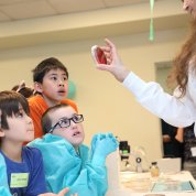 Kids in blue scrubs look in wonder at a petri dish filled with red-colored infectious bacteria.