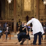 On the Cathedral stage, a seated nun receives an injection from a health worker in a lab coat.