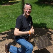 Bruce Lee gives thumbs up as he kneels, smiling next to the tree and memorial plaque honoring his late son, on the south lawn of the Clinical Center.