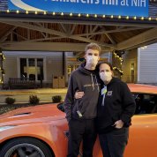 Luke and his mother Jen, wearing masks, pose in front of orange sportscar in front of the Children's Inn.