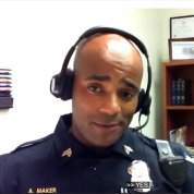 screenshot Black male police officer wearing uniform and headset, speaks into computer screen
