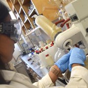 A female trainee wearing safety goggles and blue gloves does an experiment at a lab workstation, handling liquid over a large bowl. 
