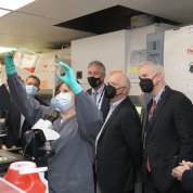 Woman in mask holds up glass flasks in gloved hands as masked people look on