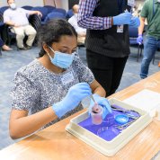 Ambati , masked, gloved and seated at table with science tray and cup in front of her.