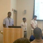 Closer audience view of Johnson at podium with other interns beside him and slide projected on screen in background.