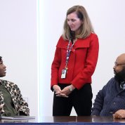 A woman standing talks to a woman and a man seated at a conference table.