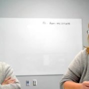 screenshot of two people at a conference table with whiteboard behind them.