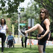 Fitness instructor does a knee lift as a woman follows and man in wheelchair looks on; orange cones behind mark route
