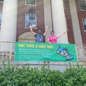 Castro and Younsi stand with arms high in the air in front of NIH Take a Hike Day banner outside Bldg. 1