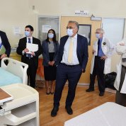 Several individuals in face masks look around a specially equipped hospital room. All wear face masks. Two men also wear labcoats.