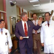 Clinton smiles and waves as he walks through a hospital corridor flanked by Fauci and Gallin
