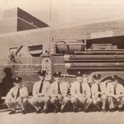 Black & white photo of firefighters sitting in front of apparatus bay.
